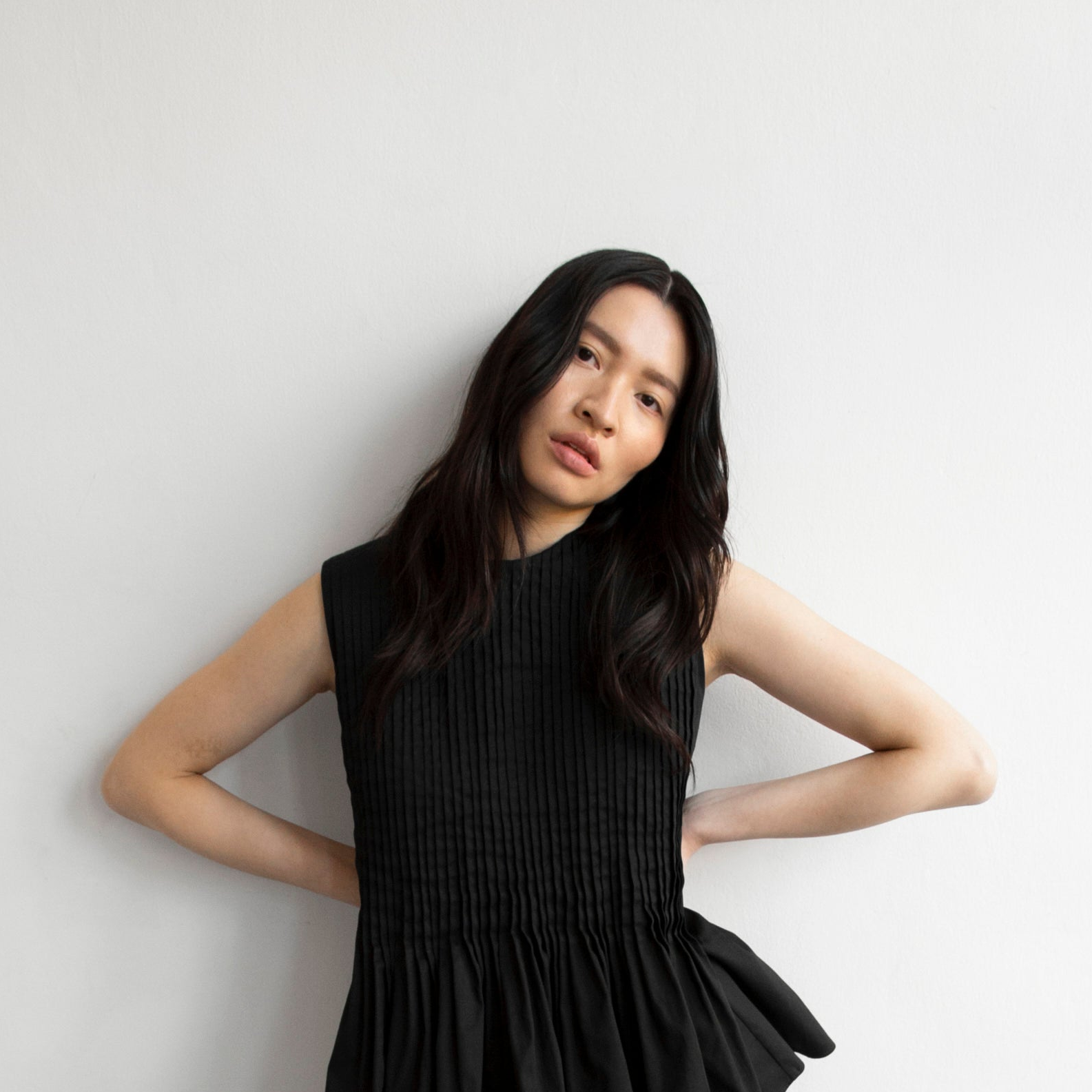 A woman with long dark hair poses in a sleeveless black pleated top with a smocked bodice and ruffled peplum hem against a plain light gray wall.